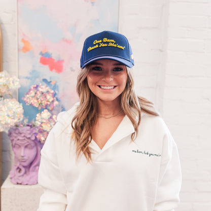 Woman wearing a blue cap with yellow text and a white sweatshirt, standing in front of a colorful abstract painting.