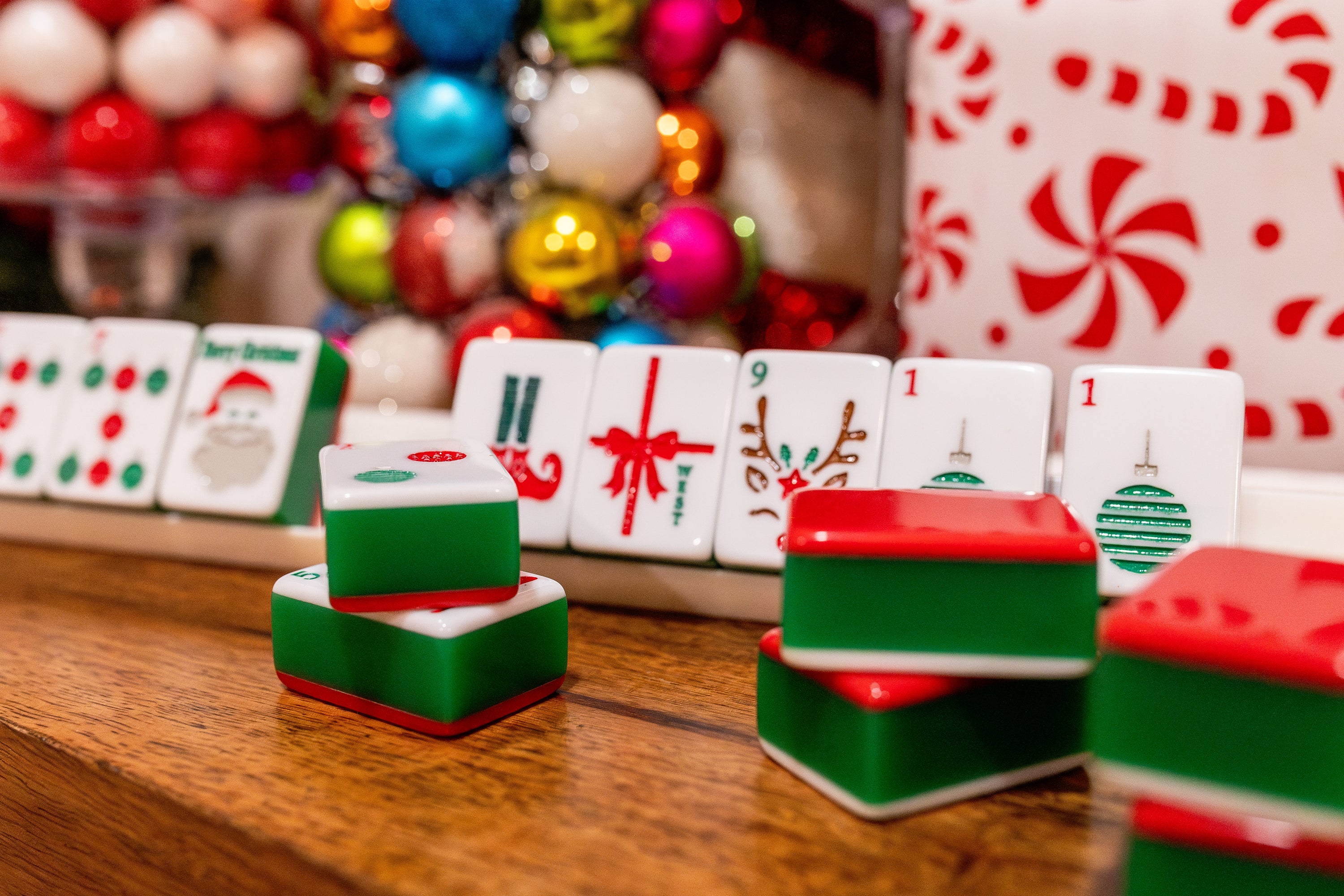 Hand-painted, multicolored mahjong tiles with festive designs, including the image of a reindeer, displayed on a table with holiday decorations in the background.